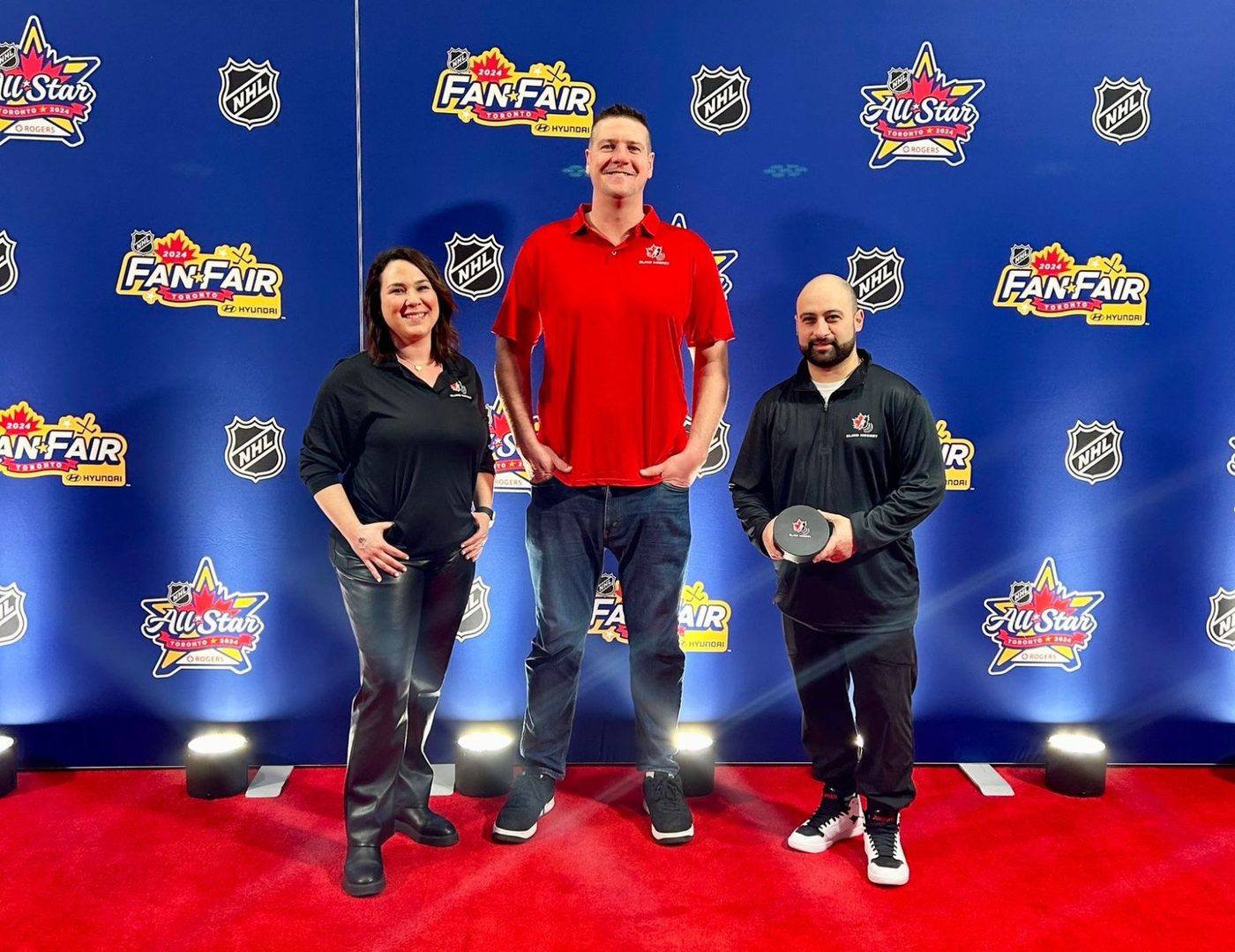 The three Canadian Blind Hockey staff members standing on a red carpet in front of a blue 2024 NHL All-Star Fan Fair backdrop, all wearing Blind Hockey shirts. The person on the right is holding the adapted Blind Hockey puck.