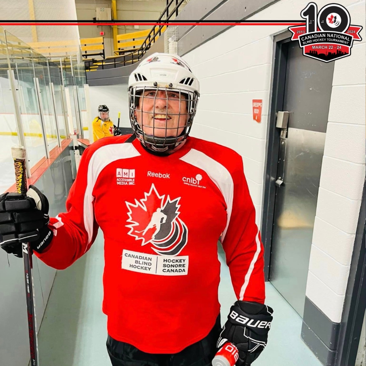 Un joueur souriant en chandail rouge de hockey sonore à l'aréna, avec le logo du 10e anniversaire du tournoi national dans le coin