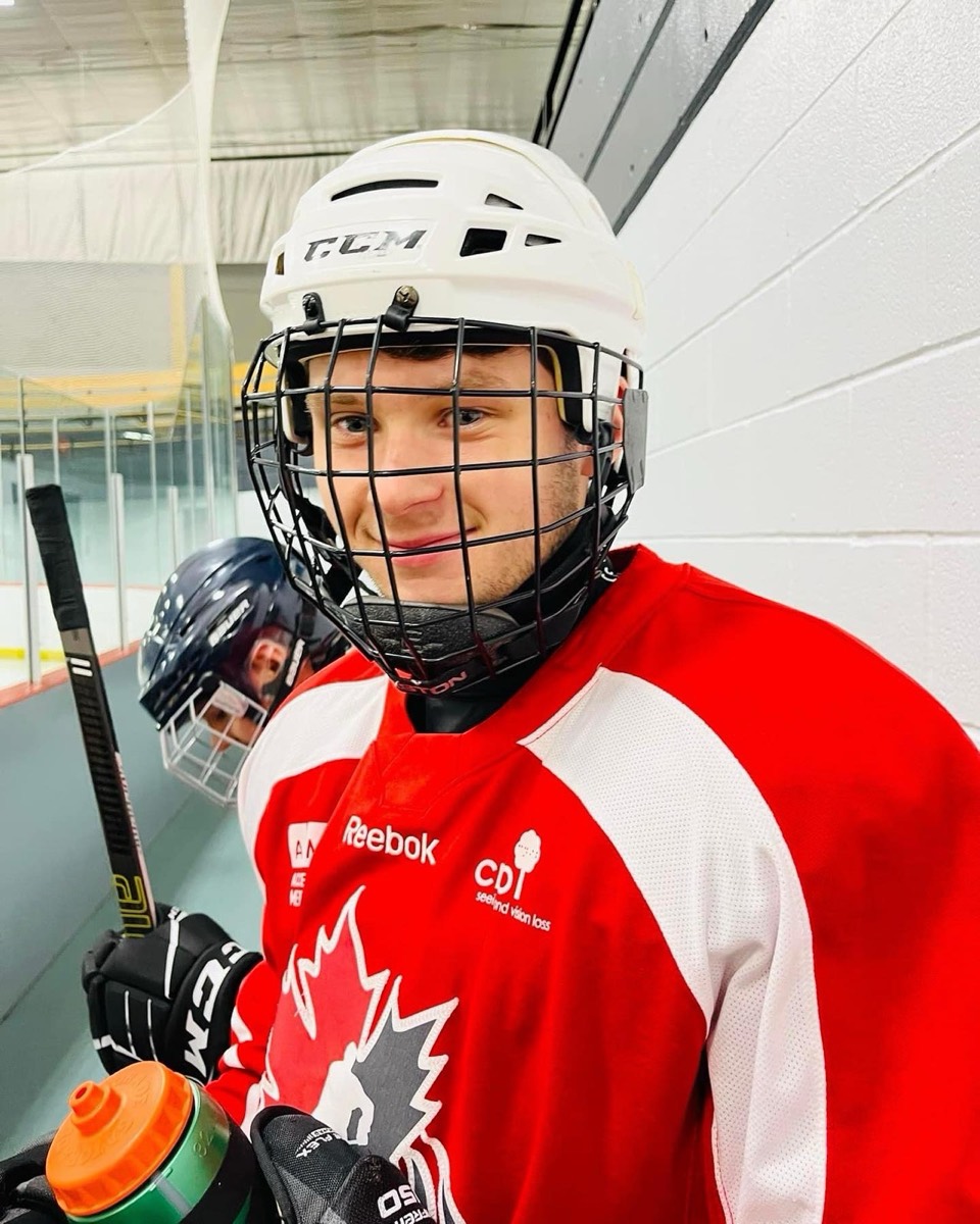 A young player in a white helmet and red Canada Blind Hockey jersey smiling at the rink boards