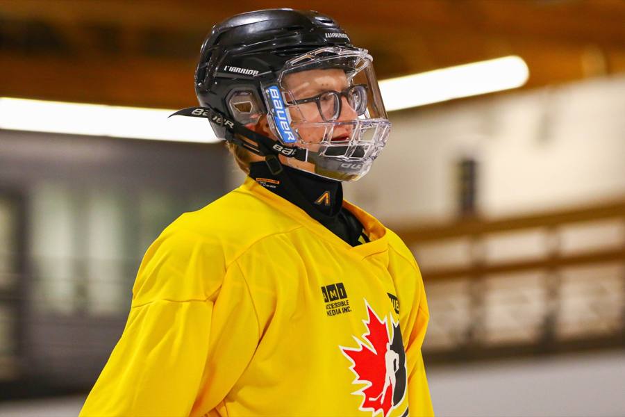 A young player in a yellow Canadian Blind Hockey jersey, glasses and a clear face shield, at the rink