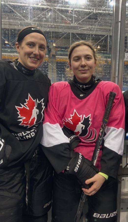 Two players smiling at the rink boards, one in a black Girls & Women's Blind Hockey jersey and one in a pink Blind Hockey jersey