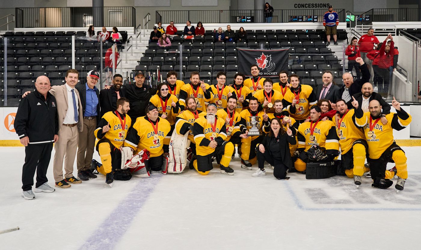 The Canadian National Blind Hockey Team in yellow Canada jerseys, wearing gold medals and gathered around the championship trophy on the ice with coaches and staff